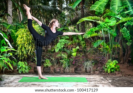 Yoga Natarajasana dancer balancing pose by woman in black cloth in the garden with palms, banana trees and plants in the pots