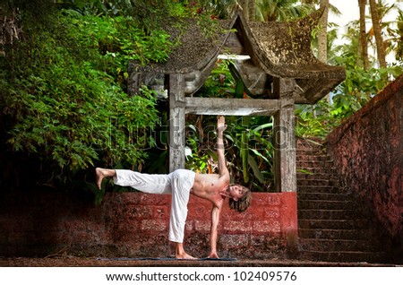 Yoga parivrtta ardha chandrasana revolved half moon pose by man in white trousers near stone temple at sunset background in tropical forest