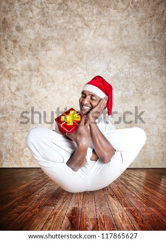 Funny Indian man in red Christmas hat doing yoga and holding red present in yoga hall with textured floor and brown background