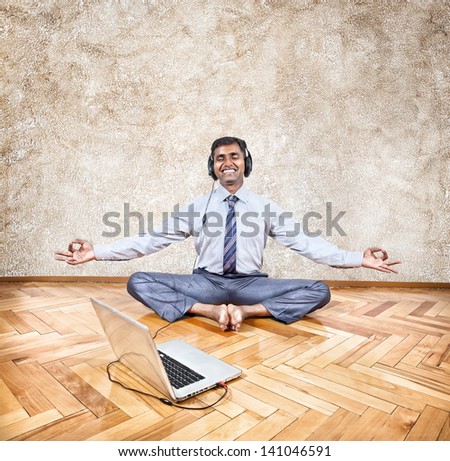Indian businessman listening to the music with headphones and doing meditation near the laptop in the office