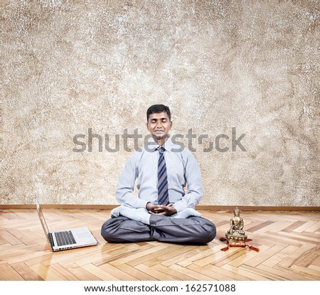 Happy Indian businessman doing meditation nearby laptop and Buddha statue in the office