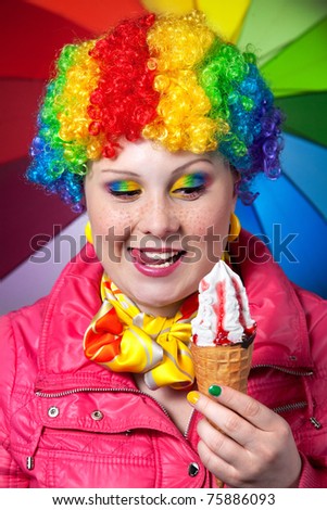 Beautiful woman in rainbow clown wig with freckles and creative rainbow make-up holding strawberry ice cream and smacks her lips at rainbow background