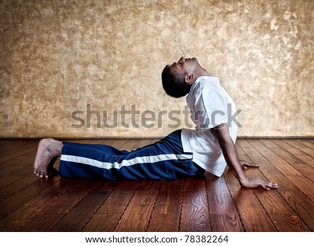 Handsome Indian man in white shirt doing bhudjangasana back bending cobra pose indoors on wooden floor at grunge background