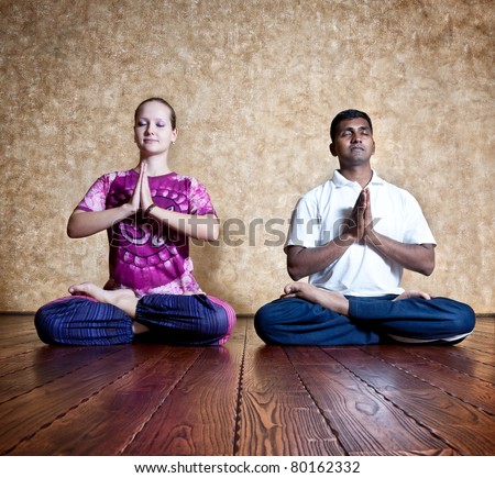 Two persons: Indian man and Caucasian woman in bright purple Indian cloth doing yoga padmasana lotus posture at the grunge background with wooden floor