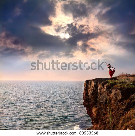 Beautiful woman doing natarajasana dancer yoga pose on the cliff near the ocean with dramatic sky at background in India