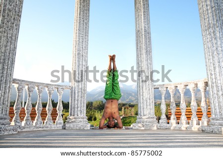 Yoga shirshasana, head standing pose by Indian man in green trousers near stone column at mountain and blue sky background