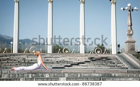 Yoga bhujangasana cobra pose by Caucasian woman in white cloth, step of surya namaskar sun salutation Exercise. Woman laying in prone position on stone stairs. Columns, mountains at background