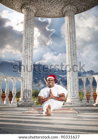 Christmas yoga eka pada shirshasana pose by happy Indian man in white costume and Christmas hat at snow mountain background.