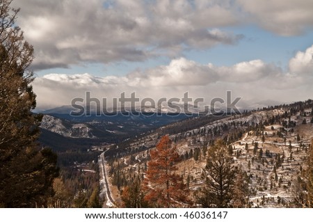 Mountain road below, snow-covered mountains in the distance, snow-covered trails, and a tree killed by pine beetles in the foreground