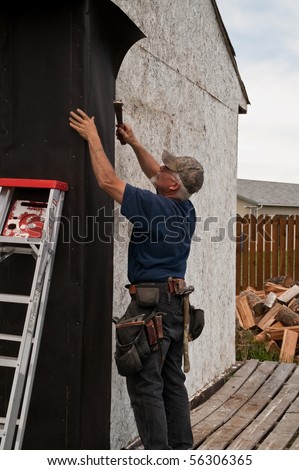 Construction worker stapling tar paper to the outside of an outbuilding prior to siding it
