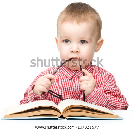 Cute little child play with book and biting glasses while sitting at table, isolated over white