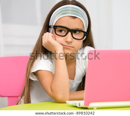 Cute little girl is sitting at table with her pink laptop and wearing glasses
