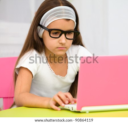 Cute little girl is sitting at table with her pink laptop and wearing glasses