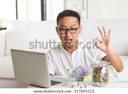 asian guy using internet computer and counting coins at home. Asian man relaxed and sitting on sofa indoor.