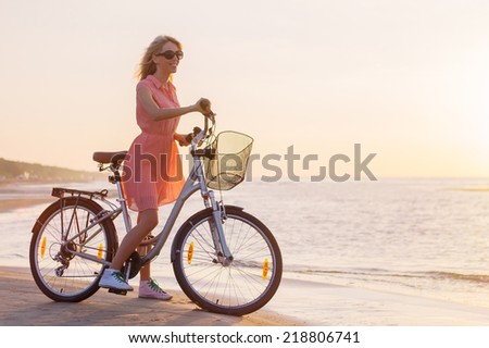 Young fashionable woman riding bicycle on the beach at sunset