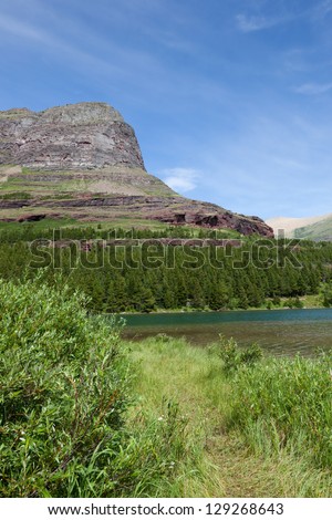 A grassy path leads into a scene of clear water green trees and the red rocky surface of a looming mountain peak in Glacier National Park, Montana.
