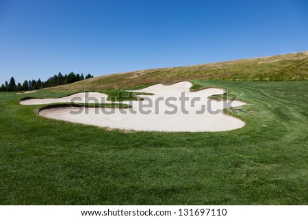 A scalloped edged sand trap with a rake in a golf course with dark green grass next to a hill with a blue sky background.
