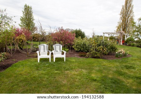 Two white lawn chairs sit next to each other in a landscaped yard with manicured grass and a background of spring bushes and trees.