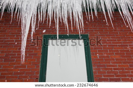 Long delicate fingers of ice hang from a gutter atop a red brick building with a green framed window reflecting the ice and snow.