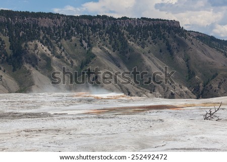 Dry mineral flats with an area of hot steam and water that drops off a ledge with distant mountains in Yellowstone National Park.