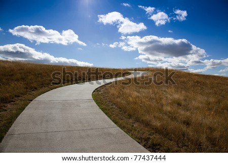 An empty golf cart path winds its way up a grassy hill towards the blue sky with fluffy white clouds and sunshine.