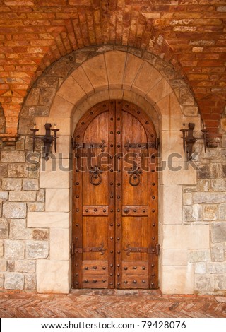 Sturdy European oak doors which are adorned by hand hammered iron accents and lamps are surrounded by masonry of chiseled stone, an arch of red brick, and a herringbone brick walkway.