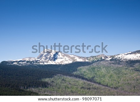 The rocky mountain of Heavens Peak still covered with snow on a bright summer day.  The foot hills below the peak show evidence of a previous wildfire leaving dead standing timber.