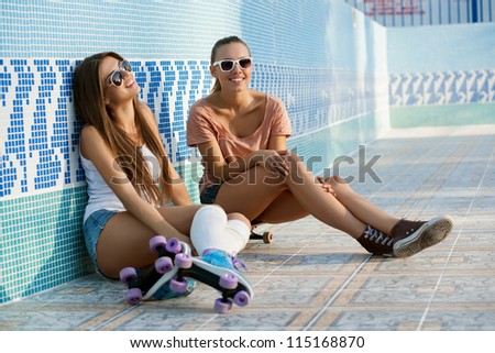 Two young skateboarding and roller skating girl friends sitting in empty swimming pool, outdoors