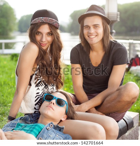 Three Happy Young People Sitting Together On Park Bench. Outdoors