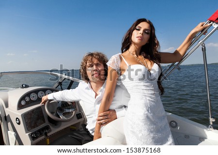 Fashion lovely happy couple posing on the luxury boat in open sea in summer. Young man and sensual brunette outdoor portrait in classic dress near the ocean. Outdoors, lifestyle.