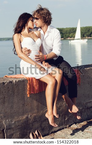 sexy lovely couple kissing in the hot sun on the pier in summer. Young man and sensual brunette outdoor portrait in classic dress near the ocean. Outdoors, lifestyle.