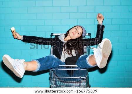 Young pretty woman holding ice cream over blue brick wall. Naughty girl having fun in shopping cart. Indoors, lifestyle
