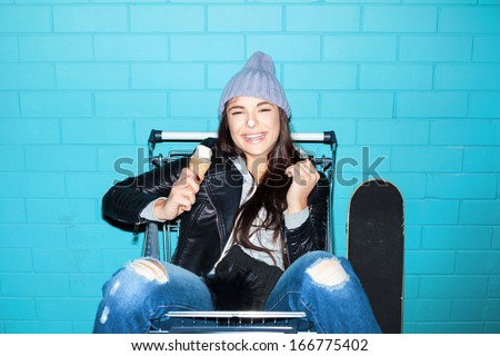 Young funky hipster woman eating ice cream over blue brick wall. Naughty girl having fun in shopping cart. Indoors, lifestyle