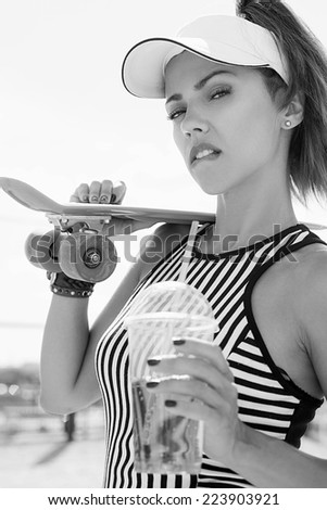 Sporty woman with skateboard drinking water against the sky. Black white portrait