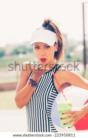 Sporty woman drinking water against the sports ground. Girl send air kiss. Outdoor lifestyle portrait