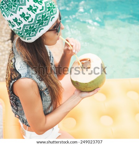 Beautiful young woman in bikini drinking coconut juice. Female legs in the pool water. Outdoor lifestyle portrait