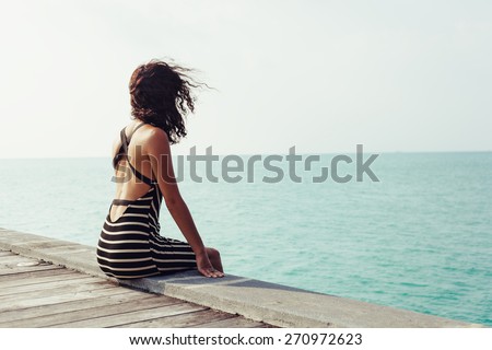 Young pretty woman sitting alone on the pier near the sea and looking on the water and waiting for somebody in ocean