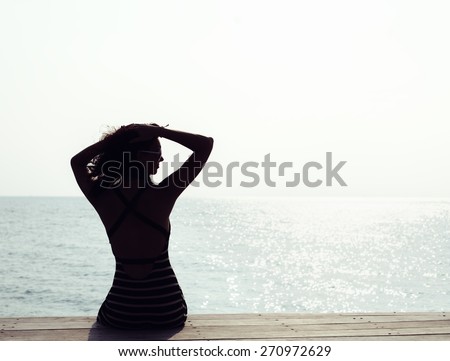Silhouette of young pretty woman sitting alone on the pier near the sea and looking on the water and waiting for somebody in ocean