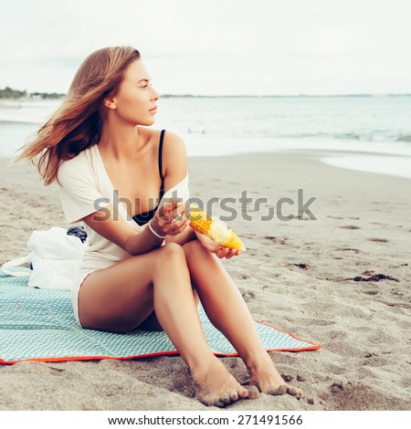 Outdoor summer style fashion portrait of young beautiful sensual woman posing on the beach on vacation. Girl holding corn and looking on the water and waiting for somebody in ocean.