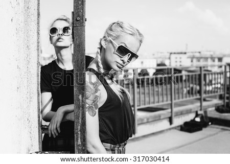 Black white portrait of two pretty blonde girls wearing plaid skirt and  black T-shirt. Girls smile, have fun against  urban city.