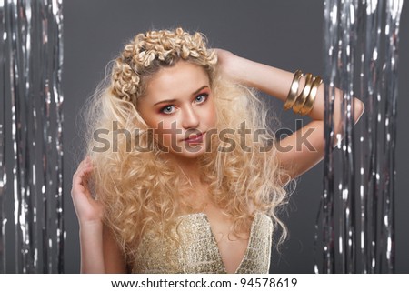 Beautiful young girl with blond curly hair, wearing golden dress and golden accessories standing behind a curtain made of silver ribbons