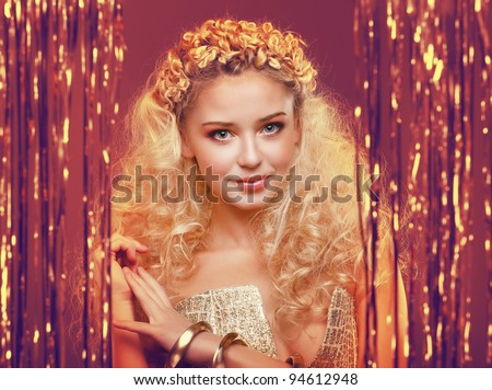 Beautiful young girl with blond curly hair, wearing golden dress and golden accessories standing behind a curtain made of golden ribbons
