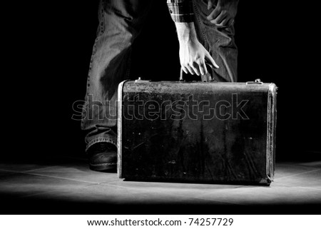 This is a high contrast, black and white image of a young man picking up an old, beat up suitcase. Shot with hard light on a black background.