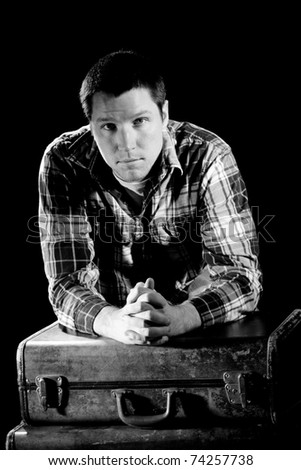 This is a high contrast, black and white image of a young man leaning on some old, beat up suitcases and looking at the camera. Shot with hard light on a black background.
