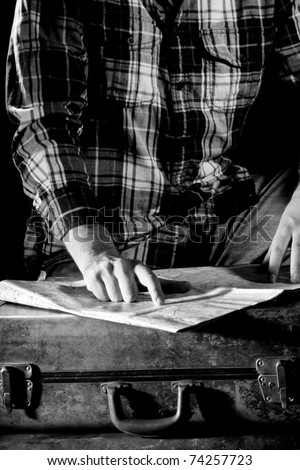 This is a high contrast, black and white image of a young man pointing on a map that is setting on top of an old, worn out suitcase. Shot with hard light.