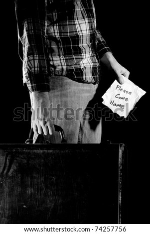 This is a high contrast, black and white image of a young man carrying an old beat up suitcase and holding a note that reads, \