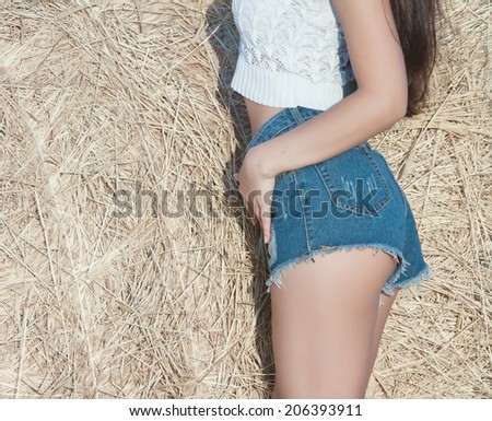 beautiful suntanned female body in short denim shorts and a white blouse on a background of haystack