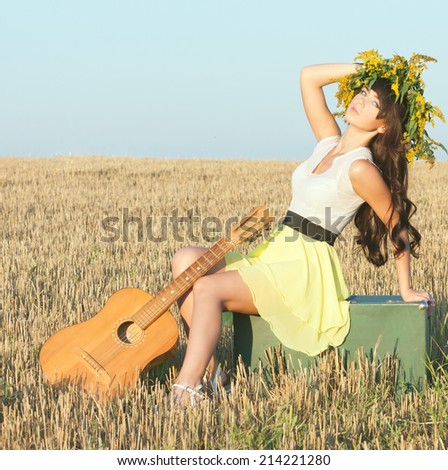 Beautiful young country girl in a short dress posing on a summer day sitting in a field on a suitcase with a guitar.Photo tinted light yellow to transfer summer atmosphere