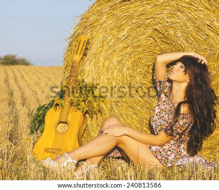 Beautiful country girl in short dress on a summer day basking in the field on a background of haystack.toned