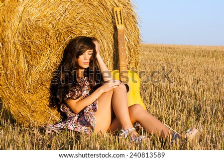 Beautiful country girl in short dress on a summer day basking in the field on a background of haystack.